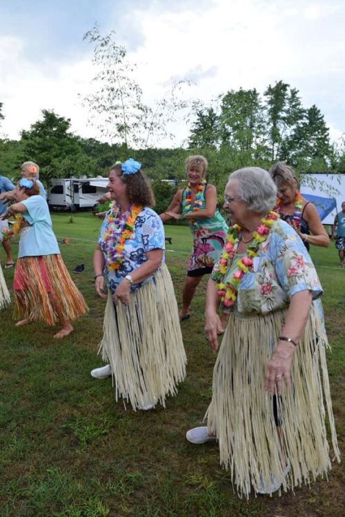 A group of women in colorful outfits and grass skirts dance joyfully outdoors, wearing flower leis and smiling.