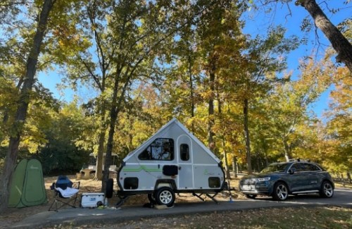 A small camper trailer parked among trees with autumn foliage, alongside a car and camping chairs.