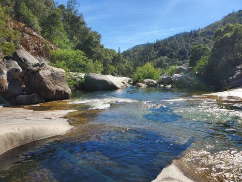 A serene river flows through rocky terrain, surrounded by lush greenery and trees under a clear blue sky.