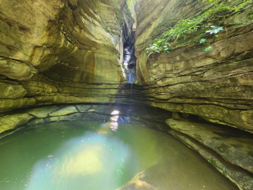 A serene canyon with smooth stone walls, a small waterfall, and a tranquil pool of water at the bottom.