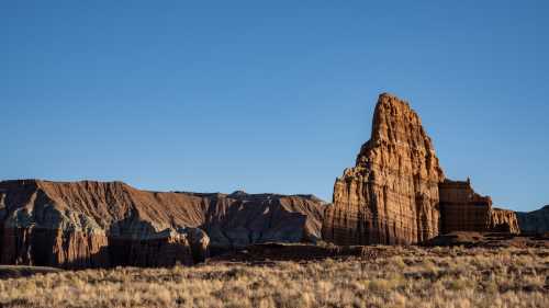 A towering rock formation rises above a desert landscape under a clear blue sky, surrounded by rugged cliffs.