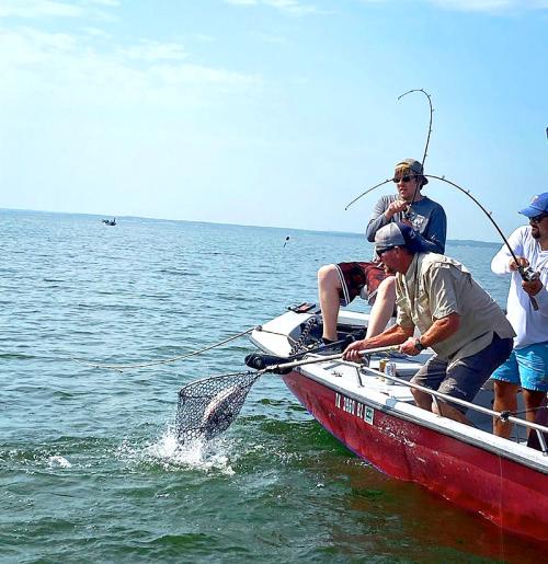 Two fishermen on a boat, one casting a line while another prepares to net a fish in calm waters.