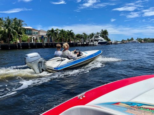 A small boat with two people speeds through a calm waterway, surrounded by palm trees and luxury homes.
