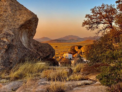 A scenic view of rocky terrain and distant mountains at sunset, with a camper nestled among the boulders.