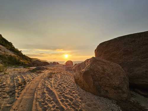 Sunset over a beach with large rocks and sandy paths, creating a serene coastal landscape.