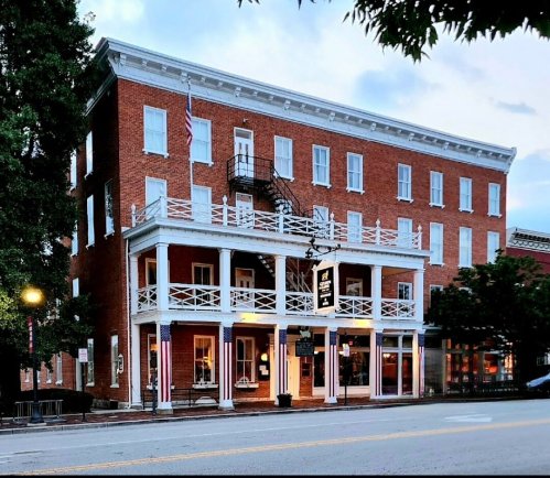 Historic brick building with a white balcony, fire escape, and American flag, set in a quaint town.