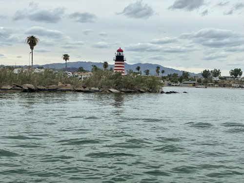 A striped lighthouse stands on a shore with palm trees, surrounded by calm water and cloudy skies in the background.