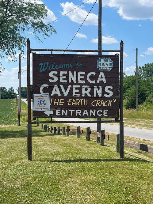 Sign welcoming visitors to Seneca Caverns, featuring "The Earth Crack" and indicating the entrance. Green fields in background.