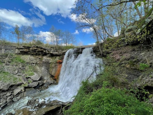 A waterfall cascades over rocky cliffs, surrounded by greenery and trees under a blue sky with fluffy clouds.