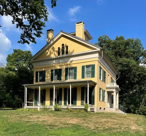 A large yellow historic house with green shutters, surrounded by trees and a clear blue sky.
