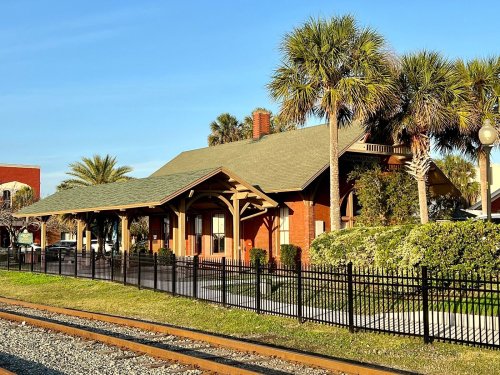 Historic train station with a green roof, surrounded by palm trees and a black fence, near railway tracks.