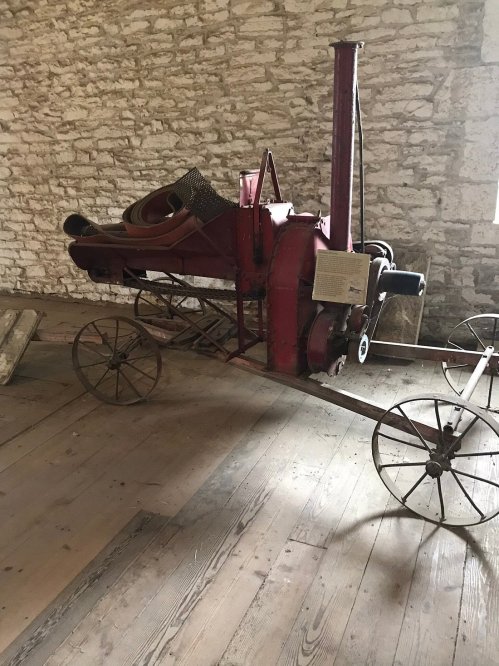 A vintage red agricultural machine on wheels, displayed on a wooden floor against a stone wall.