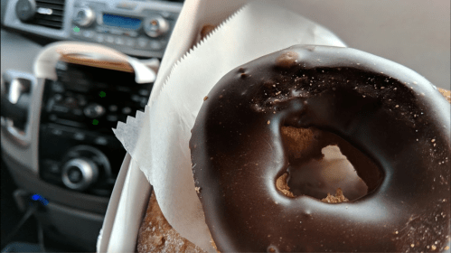 A close-up of a chocolate-glazed donut in a takeout box, with a car interior visible in the background.