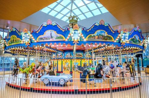 A colorful carousel featuring various animals and a car, set under a bright, glass-roofed atrium.