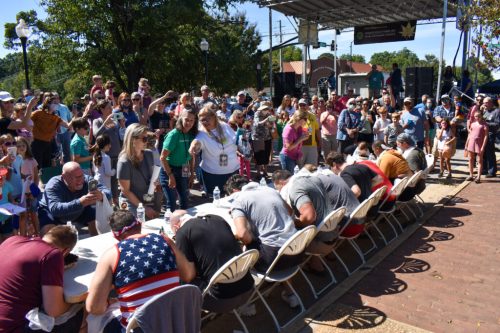 A crowd watches as participants compete in a food-eating contest at an outdoor event, with tables set up for the competitors.