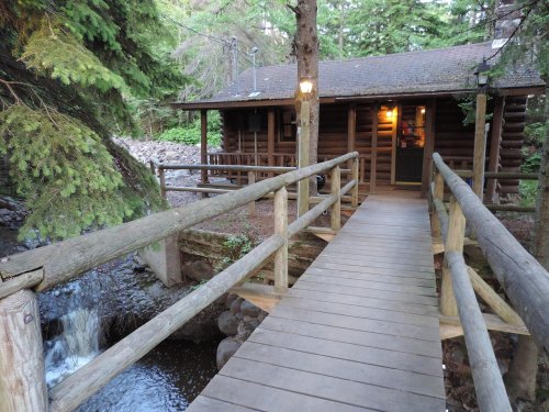A wooden bridge leads to a cozy log cabin surrounded by trees and a small stream.