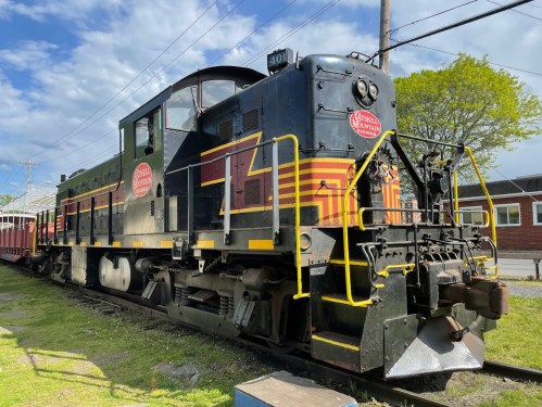 A vintage black and yellow locomotive parked on tracks, with a blue sky and green trees in the background.