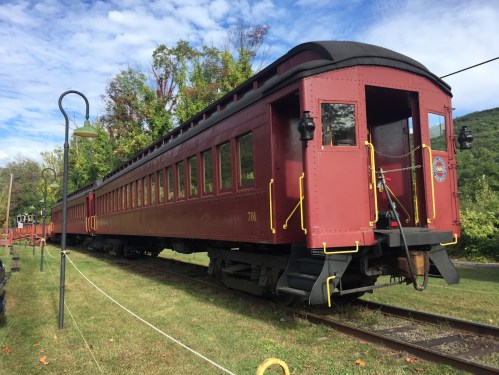 A vintage red train car parked on a grassy area, surrounded by trees and under a blue sky with scattered clouds.