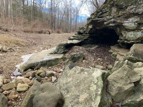 A rocky cave entrance surrounded by a dry riverbed and bare trees in a wooded area.