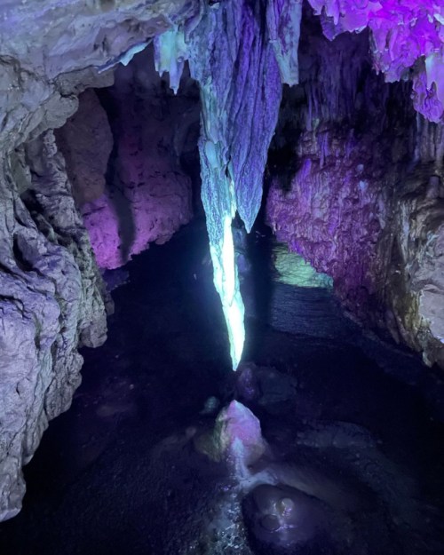 A cave interior featuring a glowing stalactite with purple lighting, reflecting in dark water below.