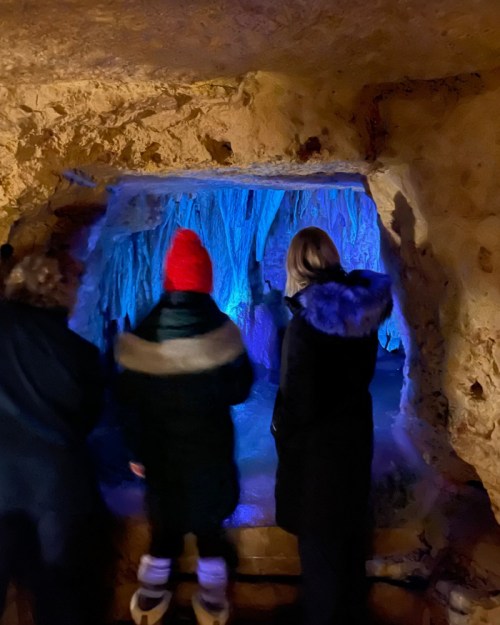 Three people stand in a cave, illuminated by blue light, gazing at icy formations.