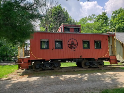 A red caboose with the B&O logo, parked on a gravel path surrounded by green trees and a blue sky.