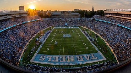 A panoramic view of a packed football stadium at sunset, with fans in blue and white cheering for the Tar Heels.