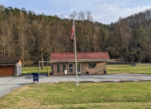 A small brick building with a red roof, flagpole, and mailbox, surrounded by trees and open grass.