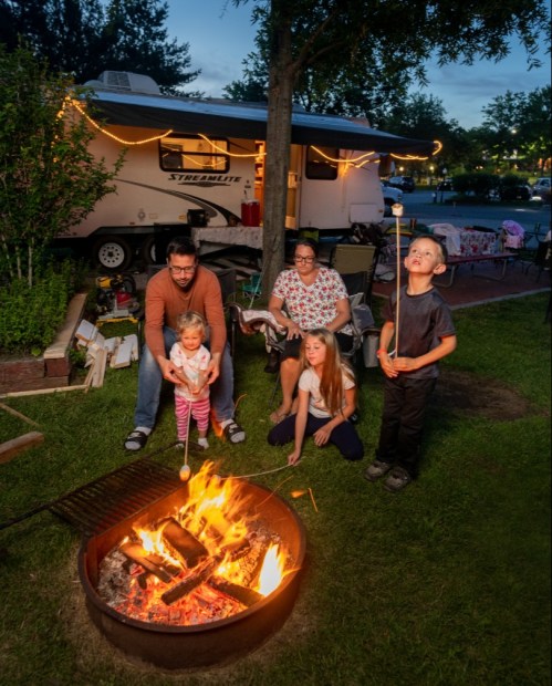 A family gathers around a campfire, roasting marshmallows, with a camper in the background and evening lights.