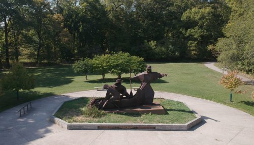 A large bronze sculpture of two figures in a park, surrounded by greenery and a walking path.