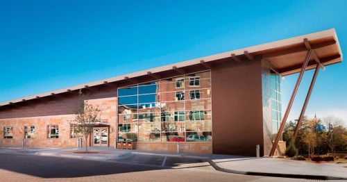 Modern building with large glass windows and a wooden overhang, reflecting the blue sky and surrounding landscape.