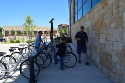 Two people near a bike rack with bicycles, one talking to a man while another sits on a bike. Bright, sunny day.