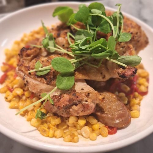 A plate of grilled chicken on a bed of corn and red peppers, garnished with fresh greens.