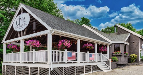 A charming building with a porch adorned with flowers and American flags, set against a blue sky with clouds.