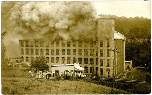 Historic photo of a factory engulfed in smoke, with a crowd gathered outside, capturing a moment of crisis.