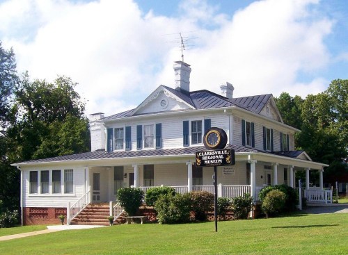 Historic white house with a black metal roof, featuring a sign for the Clarksville Regional Museum in a green landscape.