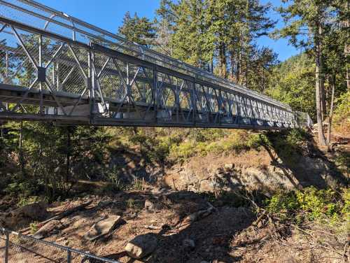 A metal pedestrian bridge spans a rocky area, surrounded by trees and greenery under a clear blue sky.