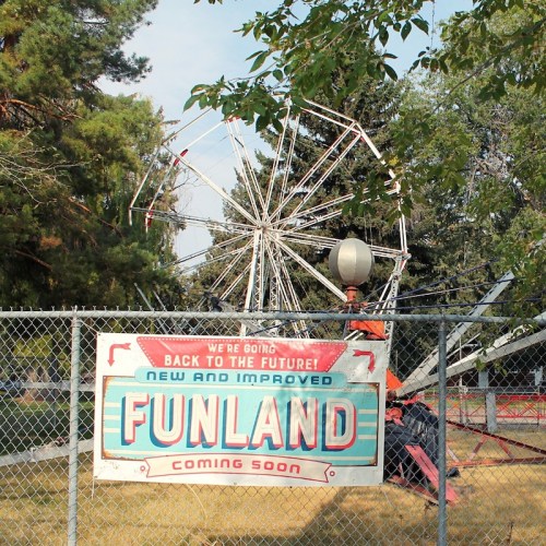 A sign reading "Funland Coming Soon" in front of a fenced amusement park ride, surrounded by trees.