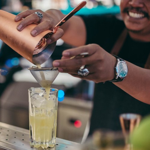 A bartender pours a cocktail through a strainer into a glass filled with ice.