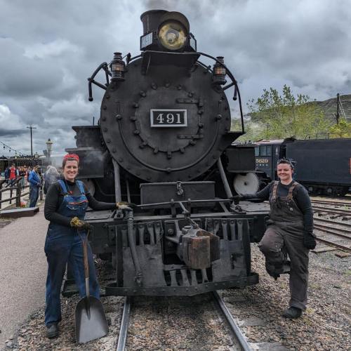Two people in work overalls stand beside a vintage steam locomotive, with a cloudy sky in the background.