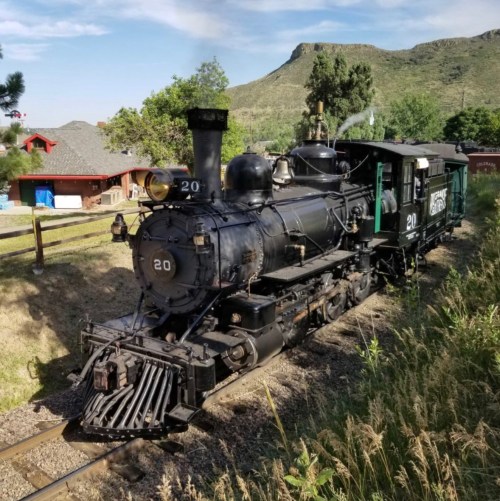 A vintage steam locomotive on tracks, surrounded by greenery and mountains in the background.