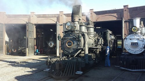 A vintage steam locomotive in a train shed, with smoke billowing and another train visible in the background.