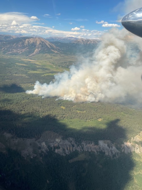 Aerial view of smoke rising from a forest fire, with mountains and green landscape in the background.