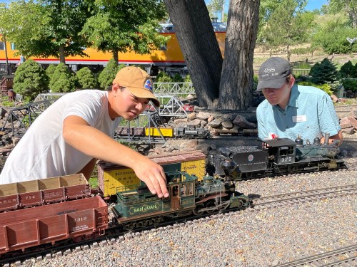 Two young men adjust model trains on a miniature railway surrounded by trees and rocks on a sunny day.