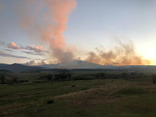 A sunset view of mountains with smoke rising in the distance, surrounded by green fields and scattered clouds.