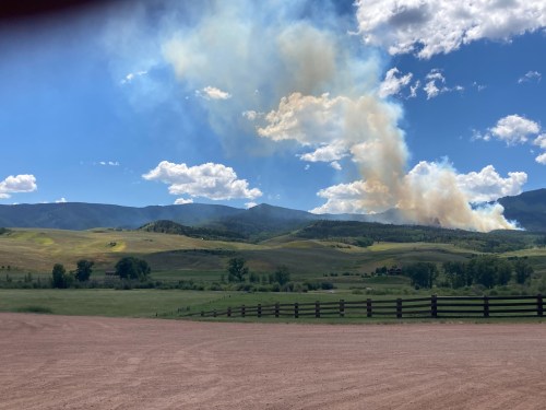 A scenic landscape with rolling hills and a plume of smoke rising against a blue sky with scattered clouds.