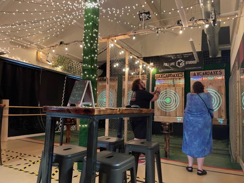 Two people throwing axes at targets in a well-lit indoor axe-throwing venue with string lights.