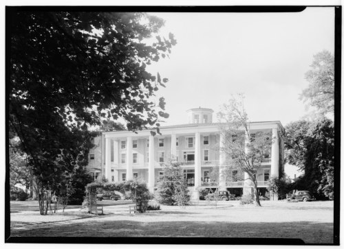 Historic building with a grand facade, surrounded by trees and vintage cars parked in front on a sunny day.