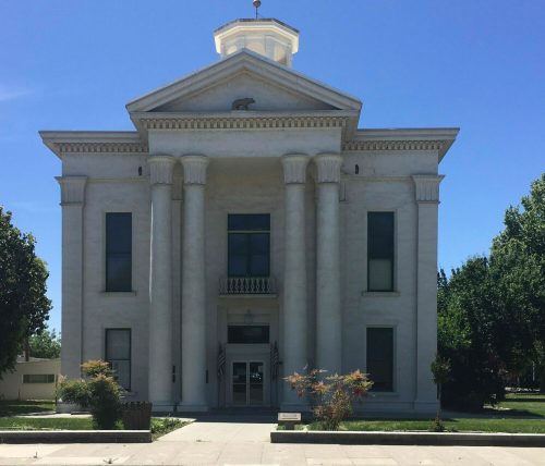 Historic white courthouse with columns, flagpoles, and a clear blue sky in the background. Green lawn in front.