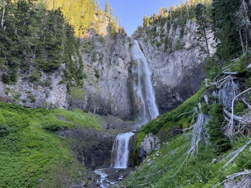 A scenic waterfall cascading down rocky cliffs, surrounded by lush greenery and tall trees under a clear blue sky.